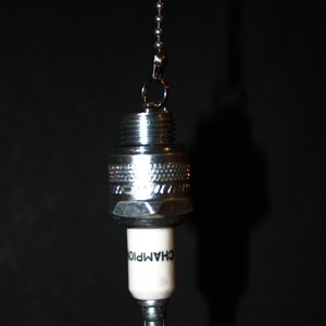 May include: A silver spark plug with a white ceramic insulator and the word "CHAMPION" printed on it. The spark plug is hanging from a silver chain.