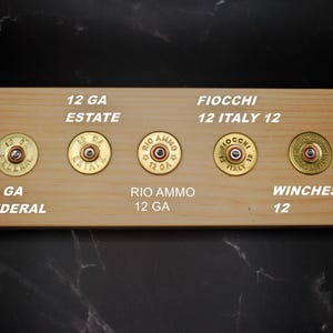 May include: A wooden display board with five 12-gauge shotgun shell casings. The casings are brass-colored with red and silver details. Text on the board includes "12 GA ESTATE", "12 GA FEDERAL", "RIO AMMO 12 GA", "FIOCCHI 12 ITALY 12", and "WINCHESTER 12".