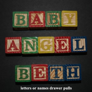 May include: Wooden alphabet blocks spelling out "BABY ANGEL BETH" in colorful letters. The blocks are arranged in three rows on a black background. The text "letters or names drawer pulls" is below the blocks.