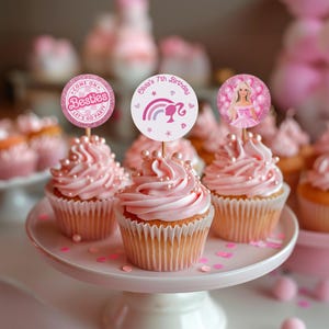 Può includere: Cupcake rosa con glassa rosa e perline su un'alzatina bianca. Ogni cupcake ha un topper decorativo con testo e immagini, tra cui un'immagine di Barbie. I cupcake sono per una festa di compleanno.