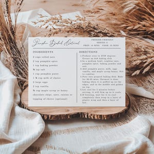 May include: A recipe card for Pumpkin Baked Oatmeal, featuring ingredients and directions. The card is on a wooden slice, surrounded by dried plants and fabric. The text includes measurements in cups and teaspoons.