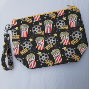 Puede incluir: Bolsa negra con cremallera con temática de cine. La bolsa tiene un patrón repetido de cubos de palomitas de maíz, carretes de película, estrellas y las palabras "Admit One". La bolsa tiene una correa negra con un clip plateado.