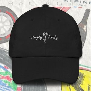 Puede incluir: Una gorra de b&eacute;isbol negra con un dise&ntilde;o bordado en blanco que dice "simply lovely" y una peque&ntilde;a flor.