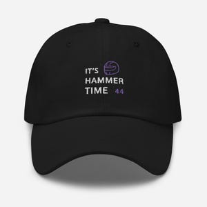 Puede incluir: Gorra de béisbol negra con el texto "IT'S HAMMER TIME 44" bordado en blanco y morado. La gorra tiene una visera curva y una corona redonda. El diseño incluye un gráfico de casco morado.