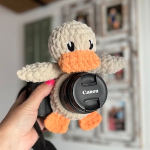 May include: A crocheted duck camera lens cover in beige and orange with black eyes. The lens cover is attached to a black camera lens with the word "Canon" printed on it.