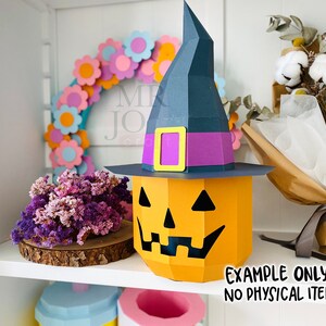 May include: A papercraft Halloween decoration featuring a jack-o'-lantern with a witch's hat. The pumpkin is orange with a carved face, and the hat is dark blue with a purple band and yellow buckle. The text "EXAMPLE ONLY NO PHYSICAL ITEM" is visible.