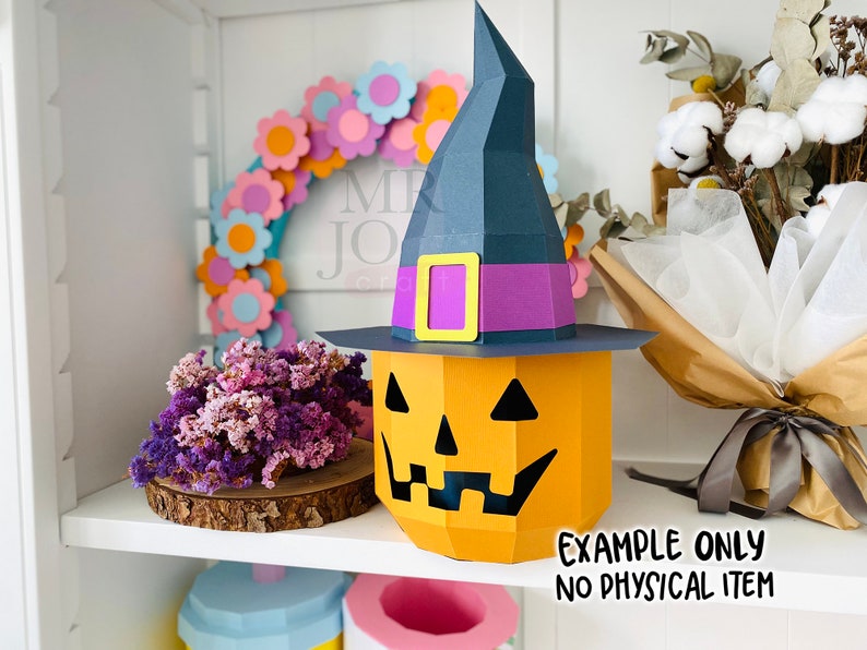 May include: A papercraft Halloween decoration featuring a jack-o'-lantern with a witch's hat. The pumpkin is orange with a carved face, and the hat is dark blue with a purple band and yellow buckle. The text "EXAMPLE ONLY NO PHYSICAL ITEM" is visible.