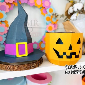 May include: A dark gray witch hat with a purple band and yellow buckle sits on a wooden slice next to a yellow jack-o'-lantern. The jack-o'-lantern has a carved face. Text reads "EXAMPLE ONLY NO PHYSICAL ITEM."