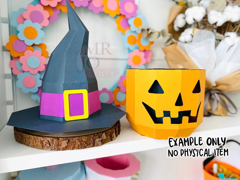 May include: A dark gray witch hat with a purple band and yellow buckle sits on a wooden slice next to a yellow jack-o'-lantern. The jack-o'-lantern has a carved face. Text reads "EXAMPLE ONLY NO PHYSICAL ITEM."