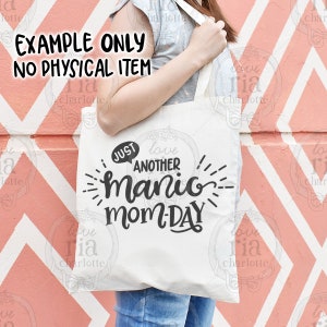 May include: A white tote bag with black text that reads "Just another manic mom-day". The text is in a playful, handwritten font. The bag is being held by a person wearing a grey shirt and blue jeans.