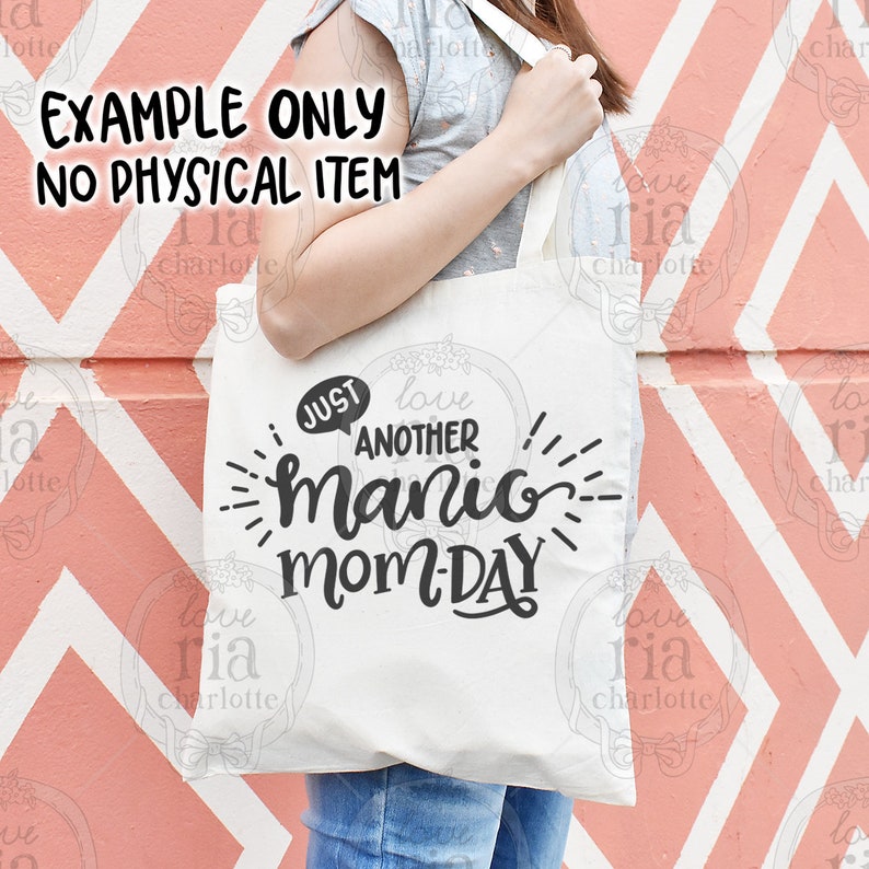 May include: A white tote bag with black text that reads "Just another manic mom-day". The text is in a playful, handwritten font. The bag is being held by a person wearing a grey shirt and blue jeans.