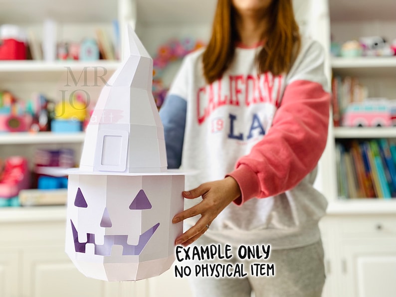 May include: A white papercraft Halloween pumpkin with a witch hat. The pumpkin has a carved face with triangular eyes and a jagged mouth. The witch hat is tall and pointed. The text "EXAMPLE ONLY NO PHYSICAL ITEM" is visible.