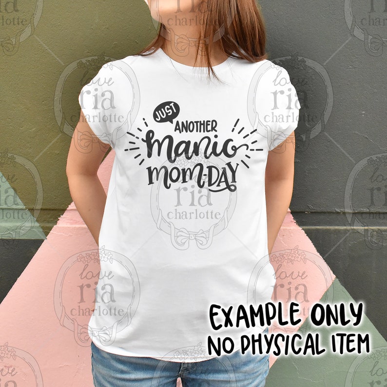 May include: White t-shirt with black text that reads "Just another manic mom-day". The text is in a playful, handwritten font. The t-shirt is being worn by a person, but only the t-shirt is visible in the image. The background is a pink and green pattern.