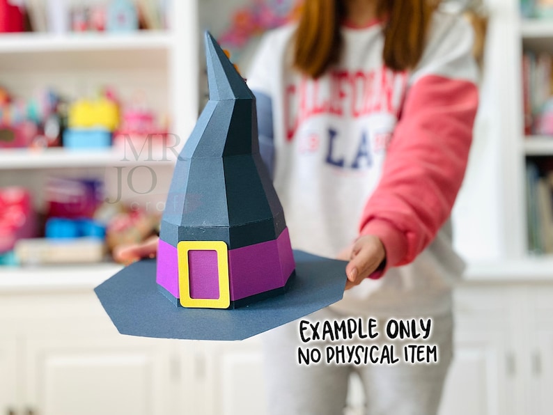 May include: A paper witch hat with a black cone and brim, a purple band, and a gold buckle. The hat is held up by a person. The text "EXAMPLE ONLY NO PHYSICAL ITEM" is printed on the bottom right.