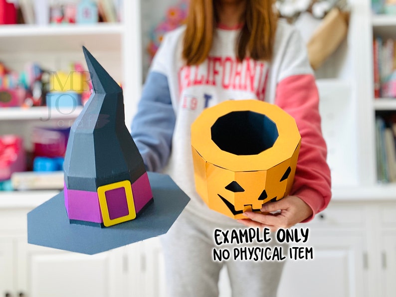 May include: A paper witch hat with a purple band and yellow buckle, and a paper jack-o'-lantern. The witch hat is dark gray and the jack-o'-lantern is orange. The text "EXAMPLE ONLY NO PHYSICAL ITEM" is visible.