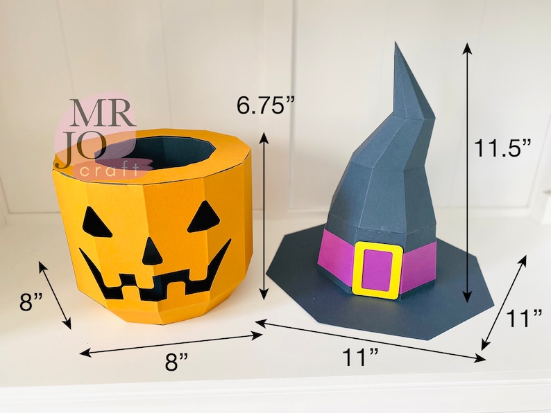 May include: A Halloween-themed papercraft set featuring a jack-o'-lantern bucket (8" x 8" x 6.75") and a witch's hat (11" x 11" x 11.5"). The jack-o'-lantern is orange with black cut-out features. The witch's hat is dark gray with a purple band and yellow buckle.