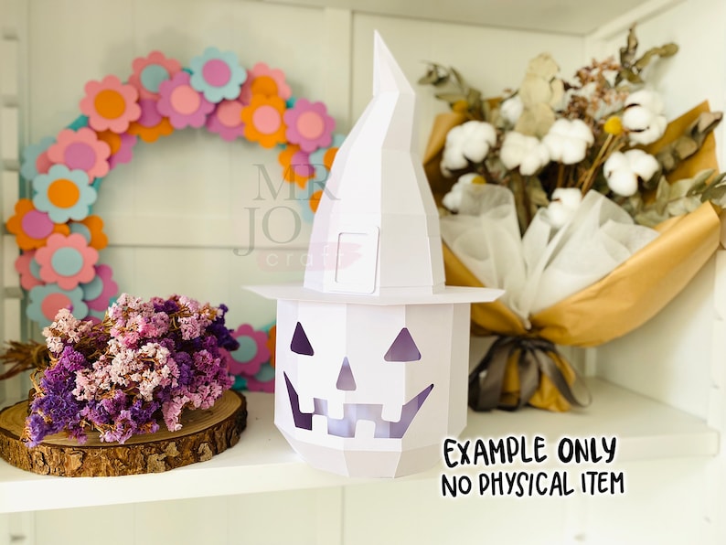 May include: A white paper jack-o'-lantern with a witch hat, a floral wreath, and dried flowers. The jack-o'-lantern has a carved face and is lit from within. The text "EXAMPLE ONLY NO PHYSICAL ITEM" is visible.
