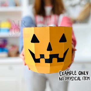 May include: A papercraft pumpkin with a jack-o'-lantern face. The pumpkin is orange with black cut-out eyes, nose, and mouth. The text "EXAMPLE ONLY NO PHYSICAL ITEM" is at the bottom. The product is held in front of a person.