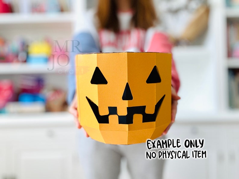 May include: A papercraft pumpkin with a jack-o'-lantern face. The pumpkin is orange with black cut-out eyes, nose, and mouth. The text "EXAMPLE ONLY NO PHYSICAL ITEM" is at the bottom. The product is held in front of a person.