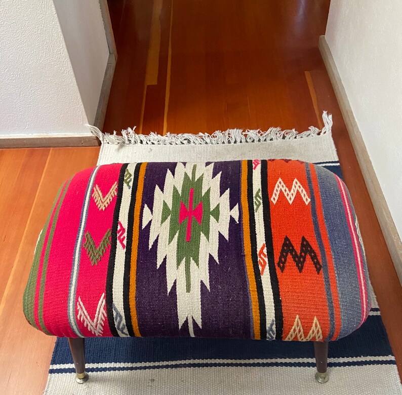 Op de afbeelding: Een kleurrijke, handgeweven ottomaan met een geometrisch patroon in tinten rood, oranje, paars en groen. De ottomaan heeft een houten frame en poten.