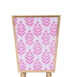 May include: A small, rectangular gold-colored metal container with a pink and white floral pattern. The container has a gold trim and sits on three small, gold-colored ball feet. The floral pattern is a repeating design of pink leaves and berries.