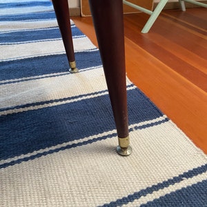 Op de afbeelding: Close-up van een houten kruk met een kleurrijke, gepatenteerde stoffen zitting en goudkleurige metalen poten. De kruk staat op een blauw-wit gestreept kleed.