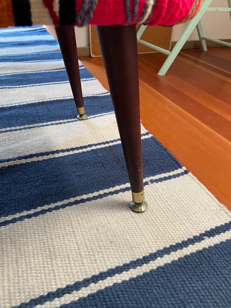 Op de afbeelding: Close-up van een houten kruk met een kleurrijke, gepatenteerde stoffen zitting en goudkleurige metalen poten. De kruk staat op een blauw-wit gestreept kleed.