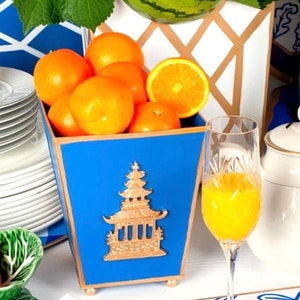 May include: A blue decorative container filled with oranges, featuring a gold pagoda design. A glass of orange juice and a white teapot are also visible. The container is part of a table setting.