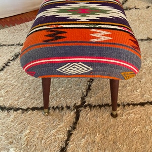 Op de afbeelding: Een kleurrijke, gepatroneerde poef met een geometrisch ontwerp in tinten roze, oranje, blauw, groen en wit. De poef heeft vier houten poten met messing puntjes en staat op een pluche, wit tapijt.