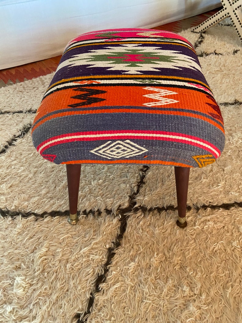 Op de afbeelding: Een kleurrijke, gepatroneerde poef met een geometrisch ontwerp in tinten roze, oranje, blauw, groen en wit. De poef heeft vier houten poten met messing puntjes en staat op een pluche, wit tapijt.