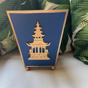 May include: A navy blue decorative box with a gold pagoda design. The box has a gold trim and sits on small gold feet. The background includes green and white tropical leaf patterned pillows.