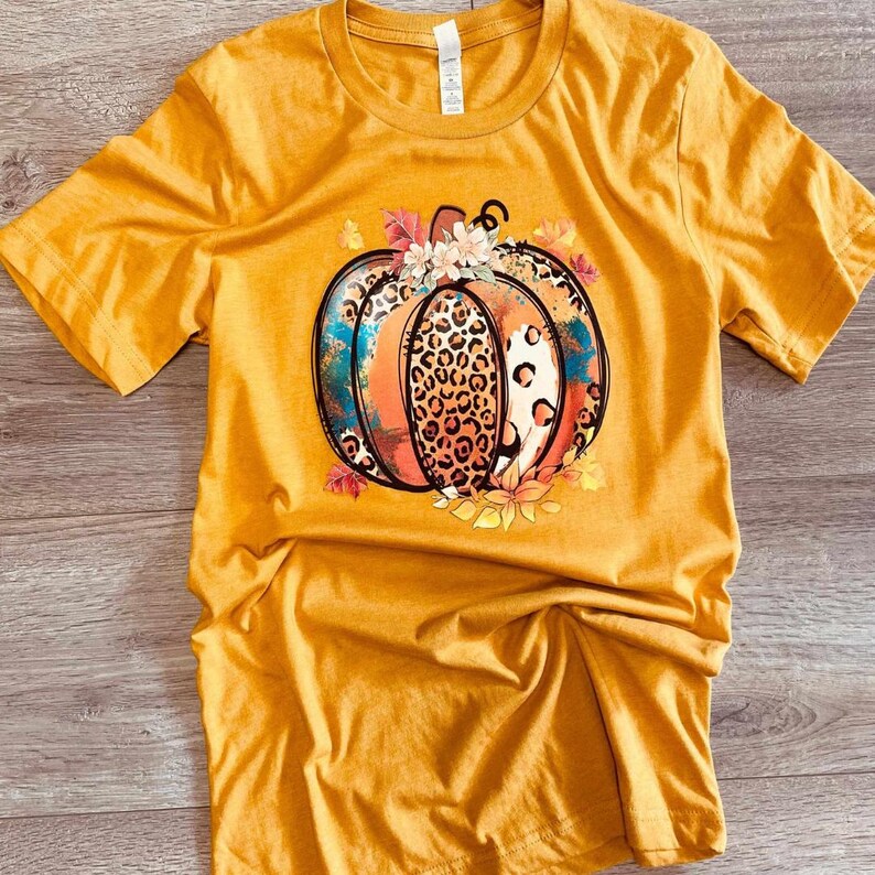 May include: A mustard yellow t-shirt with a graphic of a pumpkin with a leopard print design and fall leaves.