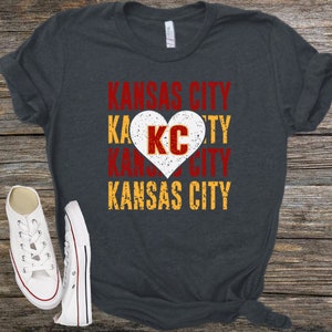 May include: A dark gray t-shirt with the words "Kansas City" in red, yellow, and white lettering. A white heart with the letters "KC" in red is in the center of the shirt.