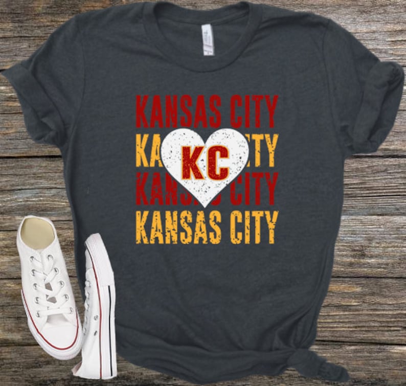 May include: A dark gray t-shirt with the words "Kansas City" in red, yellow, and white lettering. A white heart with the letters "KC" in red is in the center of the shirt.