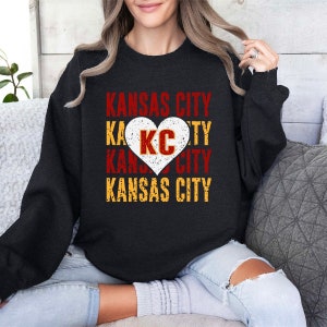 May include: Black sweatshirt with a distressed graphic design featuring the text "Kansas City" and a heart with the letters "KC" inside. The heart is red with white lettering and a distressed yellow outline.