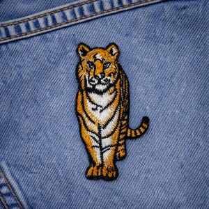 May include: Embroidered tiger patch with orange and black stripes, and white accents. The patch is sewn onto a light blue denim fabric, showcasing the detailed stitching and design of the tiger.