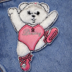 Op de afbeelding: Een witte teddybeer patch in een balletpose, met een roze turnpakje met het woord "Dancing" en roze balletschoenen. De beer heeft een zilveren tiara en staat op een blauwe denim achtergrond.