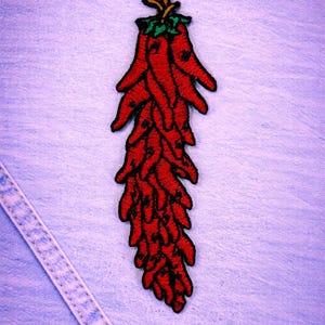 May include: Embroidered red chilli pepper patch with green and yellow detailing. The patch features a detailed design of a chilli pepper, ideal for adding a touch of spice to clothing or accessories. The patch is set against a light blue denim background.