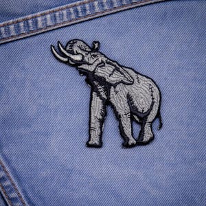 May include: Embroidered elephant patch in shades of gray and black, with detailed stitching. The patch is affixed to a light blue denim fabric, showcasing the texture of the denim and the patch's design.