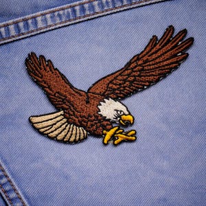 May include: Embroidered patch featuring a brown eagle in flight with outstretched wings. The eagle has a white head, yellow beak and talons, and tan wing accents. The patch is sewn onto a light blue denim fabric.
