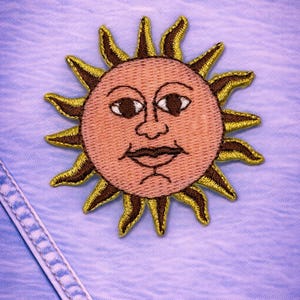 May include: Embroidered sun patch with a smiling face, featuring a peach-colored center and brown and gold rays. The patch is sewn onto a light blue denim fabric, with visible stitching.