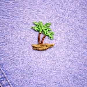 May include: An embroidered patch featuring a tropical island scene with two palm trees. The palm trees have green fronds and brown trunks, set on a tan island. The patch is on a light blue denim background.
