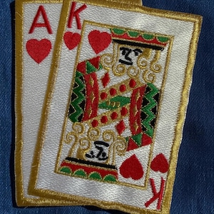 May include: Two embroidered patches shaped like playing cards, featuring the Ace of Hearts and the King of Hearts. The cards have white backgrounds, gold borders, and red heart symbols. The King figure is detailed with red, green, and black thread.
