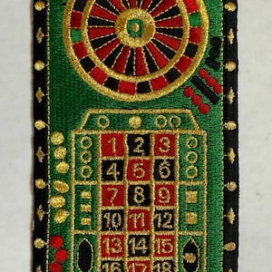 May include: A rectangular embroidered patch featuring a roulette wheel and a betting grid with numbers 1 through 21 in red, black, and green thread. The patch has a black border with gold accents and is placed above a ruler showing 3 inches.
