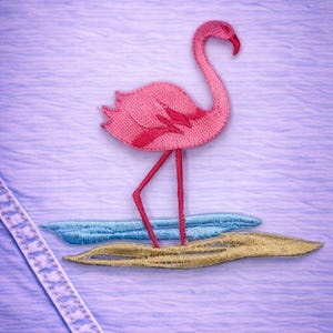 May include: Embroidered pink flamingo patch with red accents, standing on a sandy beach with blue water. The patch is sewn onto a light blue fabric, with a white stitched seam visible on the left side. A tropical theme.
