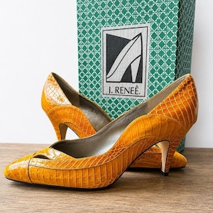 May include: A pair of mustard yellow pumps with a textured crocodile-embossed finish, featuring a pointed toe with a cutout detail and a mid-height heel. The shoes are displayed in front of a green and white patterned box labeled J. RENEÉ.