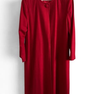 May include: A long, solid red coat featuring a round neckline, long sleeves, and a single large, matching red button closure at the top. The coat has a straight, structured silhouette and hangs from a white hanger.
