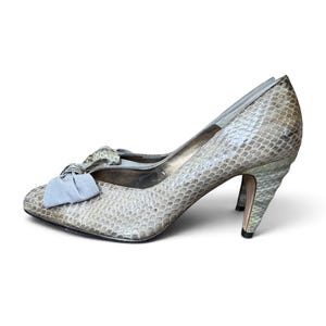May include: A pair of high-heeled pumps featuring a textured snakeskin pattern in shades of beige and gray. The shoes have a pointed toe, a mid-height heel, and a decorative light gray fabric bow attached to the front of each shoe.