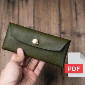 Könnte beinhalten: Eine grüne Lederbörse mit einem Messingknopfverschluss. Die Börse wird in einer Hand gehalten. PDF.
