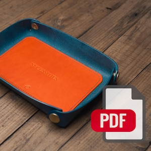 May include: A rectangular valet tray featuring a deep teal exterior and a vibrant orange interior base. The orange base is embossed with the word NECESSITIES and a small logo. The corners are secured with metallic gold-toned snap fasteners.